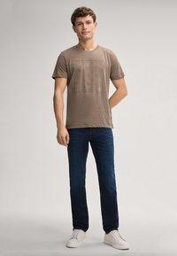 Bruine katoenen t-shirt met een grafische print in een vierkant ontwerp, gecombineerd met donkerblauwe jeans en witte sneakers. Simpele, relaxte pasvorm.