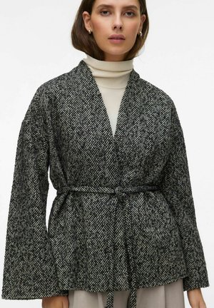 Femme portant un manteau ceinturé à motif chevrons noir et blanc par-dessus un pull à col roulé crème sur fond uni.