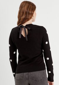 Zwarte sweater met witte stippen op de mouwen, met een sleutelgat sluiting aan de achterkant die is gestrikt met een zwarte lint. Soepele textuur, aansluitend ontwerp.