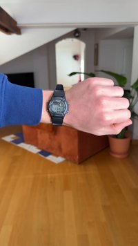 Digitale Armbanduhr mit schwarzem Gummiband, rundem Zifferblatt und grauem Display, das die Uhrzeit anzeigt. Verfügt über Tasten an der Seite und eine strukturierte Oberfläche.