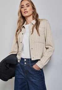Beige randig knappjacka med en kortad design, ihopparad med en vit topp och mörkblå jeans. Har en fungerande ficka och tryckknappar.