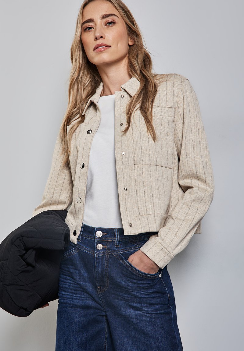 Beige randig knappjacka med en kortad design, ihopparad med en vit topp och mörkblå jeans. Har en fungerande ficka och tryckknappar.