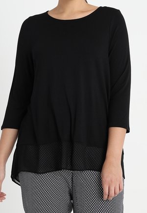 Schwarzes Dreiviertelarm-Oberteil mit Netzsaum, getragen zu schwarz-weiß gemusterten Hosen an einem Model vor weißem Hintergrund.