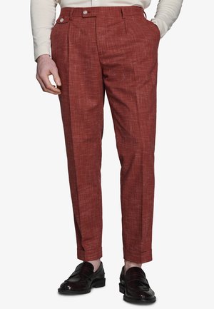 Uomo che indossa pantaloni eleganti in tessuto color ruggine e mocassini neri in pelle, con una mano in tasca e camicia bianca infilata.