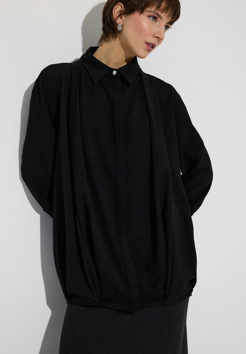 Γυναίκα που φοράει υπερμεγέθη μαύρο πουκάμισο με κουμπιά και γιακά, στεκόμενη μπροστά σε λευκό τοίχο, κοιτάζει προς τα δεξιά, με τα χέρια πίσω από την πλάτη.