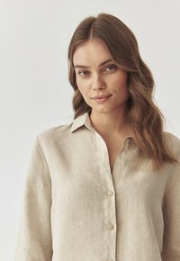 Vaalean beige pellavapaita, jossa on napitus eteen, kaulus ja pitkät hihat. Kevyt texture ja rento istuvuus.