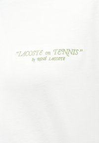 Λευκό ύφασμα με κεντημένο κείμενο σε ανοιχτό πράσινο που λέει «LACOSTE on TENNIS» από τον René Lacoste σε γραμμματοσειρά χειρόγραφου.