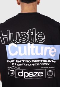Crna pamučna majica s velikim bijelim i svijetloplavim natpisom na leđima, s frazama poput "Hustle Culture" i detaljima brenda.