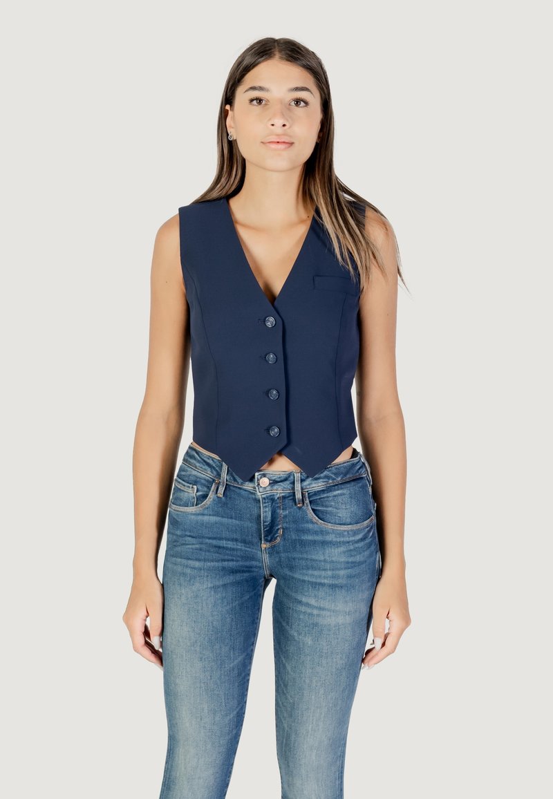 Gilet marine avec un décolleté en V profond, cinq boutons et une poche sur la poitrine à gauche ; associé à un jean skinny bleu clair, au design ajusté.