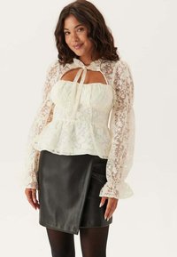 Witte kanten blouse met opbollende doorschijnende mouwen, voorzien van een tailleband en hartvormige halslijn, gecombineerd met een zwarte leren wikkelrok.
