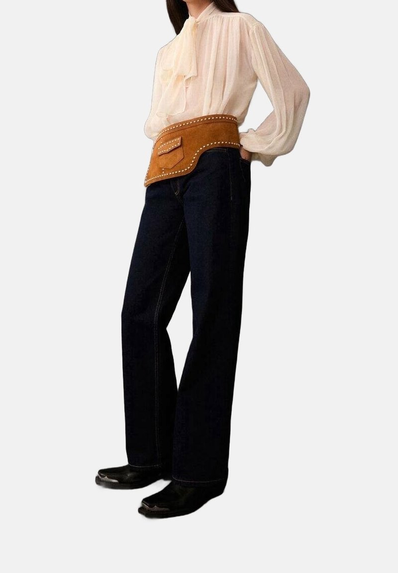 Personne portant un chemisier blanc transparent à manches longues, un jean sombre à jambes larges, des bottes noires et une grande pochette ceinture en cuir marron cloutée.