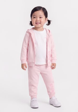 Lächelndes junges Mädchen mit Zöpfen, das einen hellrosa Hoodie und Hose mit weißen Sneakers vor weißem Hintergrund trägt.