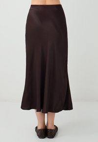Jupe maxi en satin marron avec une forme évasée, une texture lisse et une taille ajustée, associée à des chaussures tissées marron.