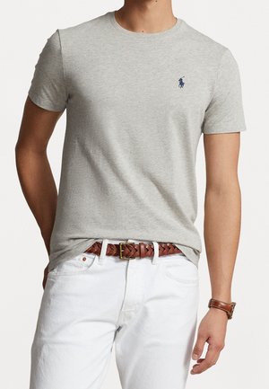 Graues Baumwoll-T-Shirt mit Rundhalsausschnitt und kurzen Ärmeln; verfügt über ein kleines blaues Logo auf der linken Brust. Getragen mit weißen Hosen und einem geflochtenen braunen Gürtel.