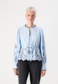 Blusa azzurro chiaro con maniche lunghe, colletto arricciato, chiusura con bottoni e motivi floreali ricamati lungo l'orlo e le maniche.