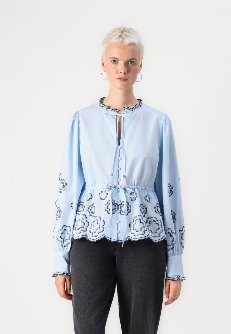 Blusa azzurro chiaro con maniche lunghe, colletto arricciato, chiusura con bottoni e motivi floreali ricamati lungo l'orlo e le maniche.