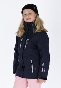 Granatowa zimowa kurtka z wysokim kołnierzem, zapinanymi kieszeniami, teksturowanym materiałem i logo. Noszona z jasnoróżowymi spodniami i czarną czapką. 