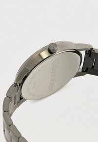 Ρολόι Calvin Klein από ανοξείδωτο ατσάλι με βραχιόλι από μεταλλικές αλυσίδες, κορώνα στο πλάι με χάραξη "CK" και λεπτομέρειες χαραγμένες στο πίσω καπάκι.