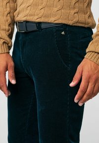 Smaragdgroene corduroybroek met verticale ribbels, voorzien van zijzakken en een klein gouden accent nabij de tailleband, gecombineerd met een beige kabelgebreide trui.