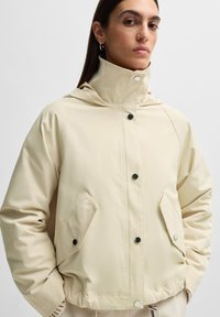 Jachetă impermeabilă bej cu glugă, butoane negre, două buzunare laterale și o croială relaxată. Textură netedă și design minimalist.