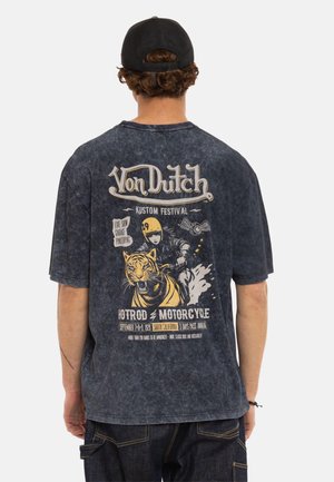 Personne portant une casquette noire et un T-shirt Von Dutch noir délavé avec un graphisme de moto et de tigre ainsi qu'un texte de festival dans le dos.