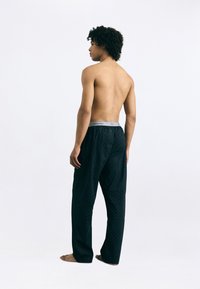 Čierne pyžamové nohavice s vzorom a šedým elastickým pásom s nápisom "Calvin Klein." Hladká textúra a uvoľnený strih. Bez trička, model otočený chrbtom.