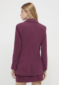 Donna con lunghi capelli biondi, che indossa un blazer di velluto a coste viola e una gonna abbinata, di spalle contro uno sfondo bianco puro.