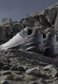 Γκρι και λευκά παπούτσια πεζοπορίας Adidas Terrex τοποθετημένα σε πετρώδη έκταση με μεγάλα βράχια στο φόντο κάτω από καθαρό ουρανό.