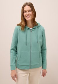 Felpa leggera verde chiaro con zip e cappuccio, tasche frontali e cordini. Presenta una texture morbida e un design dalla vestibilità comoda.