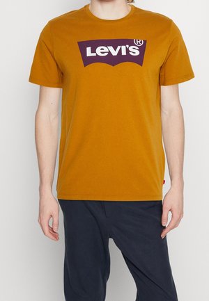 Osoba ubrana w musztardowo-żółtą koszulkę Levi's z logo w kolorze fioletowo-białym, w połączeniu z ciemnoniebieskimi spodniami, stojąca na jednolitym tle.