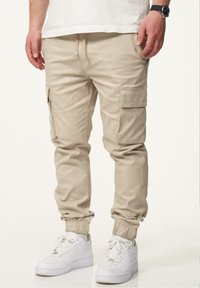 Beige Cargo-Hosen mit elastischen Bündchen, Seitentaschen und Kordelzugbund. Kombiniert mit weißen Sportschuhen für einen lässigen Look.