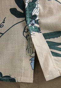 Camicia a maniche corte in tessuto beige con motivi di foglie verdi, chiusura con bottoni e etichetta tessuta. Presenta un design con sfondo strutturato.