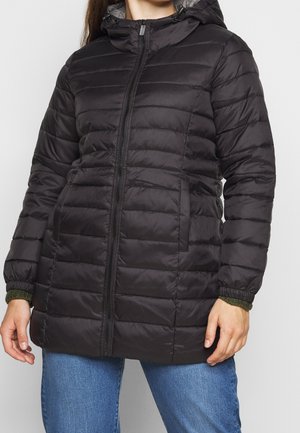Personne portant une veste matelassée noire avec capuche, fermeture éclair à l'avant et poches, associée à un jean bleu.