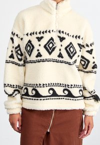 Fleecový svetr v krémové barvě s černými geometrickými vzory a lemováním. Má vysoký límec a půlzip. Měkká, strukturovaná tkanina.