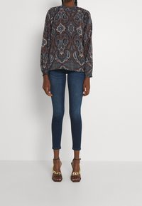 Chemisier à manches longues avec un motif paisley dans des teintes de bleu, marron et blanc, associé à un jean en denim foncé ajusté et des sandales à talons marron.