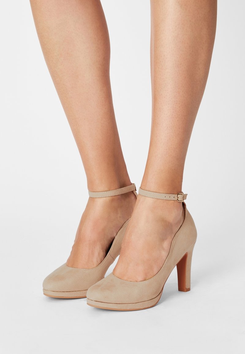 Jambes portant des chaussures à talons hauts en daim beige avec des brides à la cheville et des bouts fermés arrondis sur un fond clair uni.