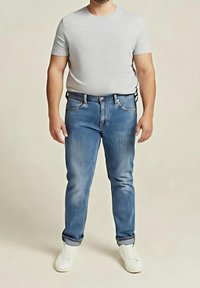 Homme portant un t-shirt gris clair à manches courtes, un jean bleu avec les revers roulés et des baskets blanches, debout devant un fond uni.
