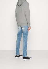 Sudadera gris con capucha combinada con jeans azules claros desgastados. Zapatillas negras de corte bajo con suelas blancas completan el atuendo.
