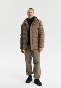 Brązowa kurtka puchowa z kapturem, zamykanymi kieszeniami i elastycznymi mankietami. W zestawie ze luźnymi beżowymi spodniami i czarnym obuwiem.