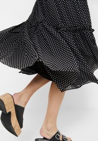 Robe noire à pois blancs tourbillonnant au-dessus de jambes nues portant des sandales à plateforme noires avec des talons en liège et deux boucles.