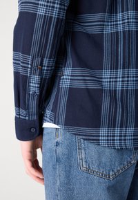 Camisa azul marinho e clara de padrão xadrez com punhos de botão, usada sobre uma camisa branca. Combinando com jeans de denim claro com costura amarela.