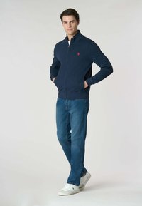 Giacca zip-up blu navy con colletto alto, abbinata a jeans azzurri e sneakers bianche. Presenta tasche laterali e un piccolo logo sul petto.