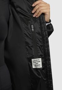 Schwarze gepolsterte Jacke mit glatter Textur, ausgestattet mit einer Reißverschlusstasche und einem Etikett, das besagt: "Eine warme Umarmung aus Stoff."