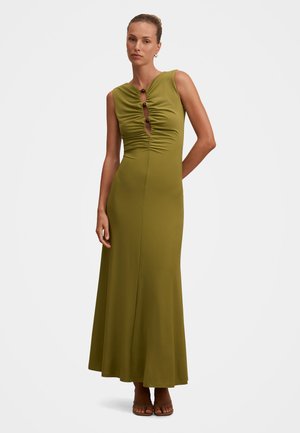 Kvinde iført en ærmeløs olivengrøn maxikjole med rynket forside og tre knapper, stående med den ene hånd bag ryggen.