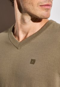 Khaki-grön V-ringad tröja av stickad tyg; har en strukturerad yta och en liten fyrkantig logotypdetalj.