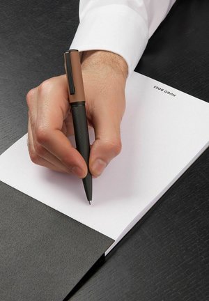 Čierne textúrované pero s hnedým viečkom držané v ruke, píše na bielom notese na tmavom povrchu. Notepad obsahuje logo.