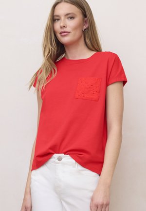 Junge Frau mit langen blonden Haaren, die ein rotes Kurzarmshirt mit einer strukturierten Brusttasche und weiße Hosen vor einem einfarbigen Hintergrund trägt.