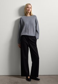 Pull en tricot gris avec un col large, associé à un pantalon noir à large jambe. Le mannequin se tient devant un fond neutre.