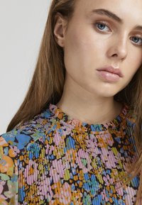 Daudzkrāsaina plisētā blūze ar noapaļotu kakla izgriezumu, ar zilu, rozā, oranžu un zaļu ziedu rakstu uz tumša fona.