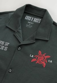 Tmavě zelené polo tričko se strukturovaným límcem, zobrazující bílé texty "GUNS N' ROSES" a grafiku červené růže s textem "LA CA" pod ní.